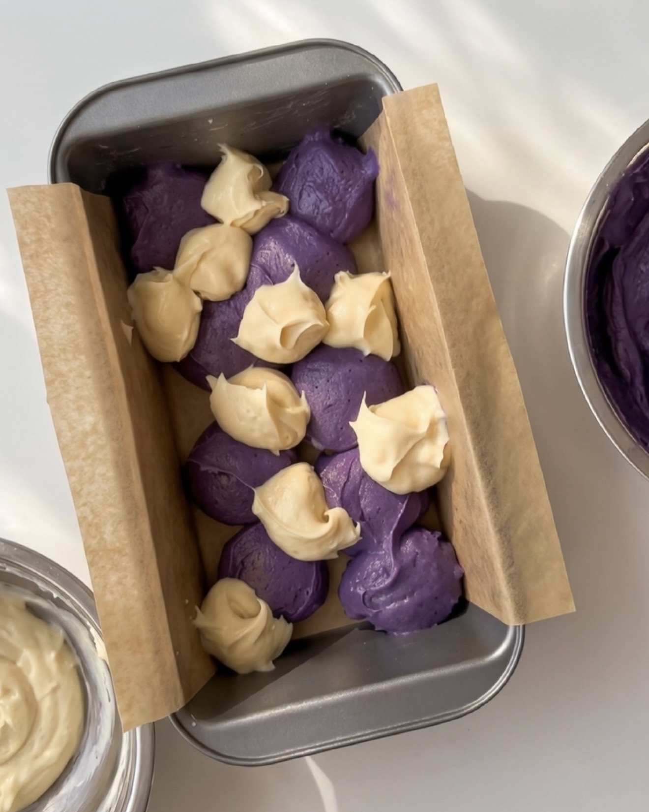 Cucharadas de masa de ube morado y masa de vainilla alternadas en un molde para pan forrado para el bizcocho mármol de ube Ube Nolia