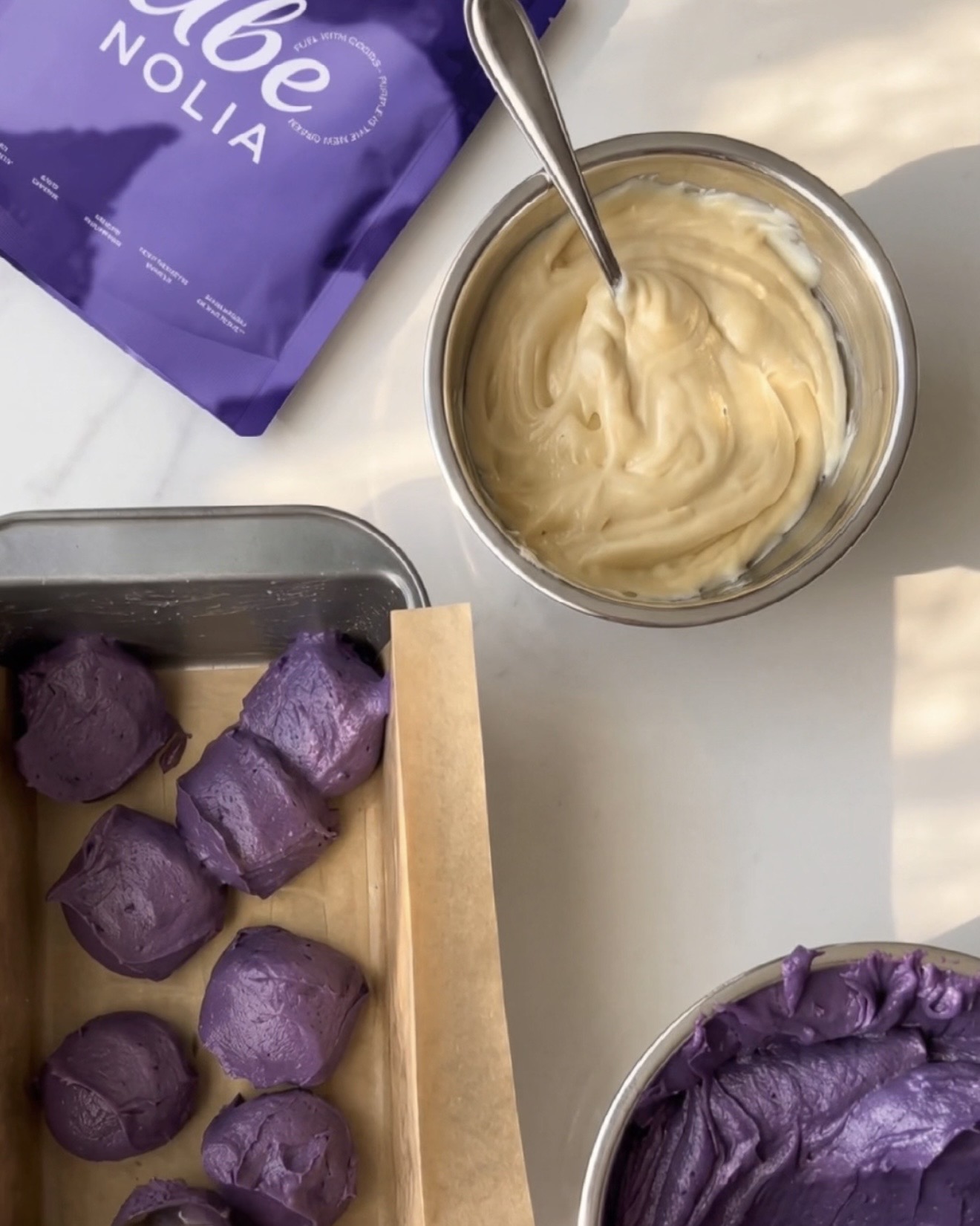 Ube halaya casero terminado, masa de vainilla y cucharadas de masa de ube morado en un molde para pan junto a una bolsa de Ube Nolia polvo de ube