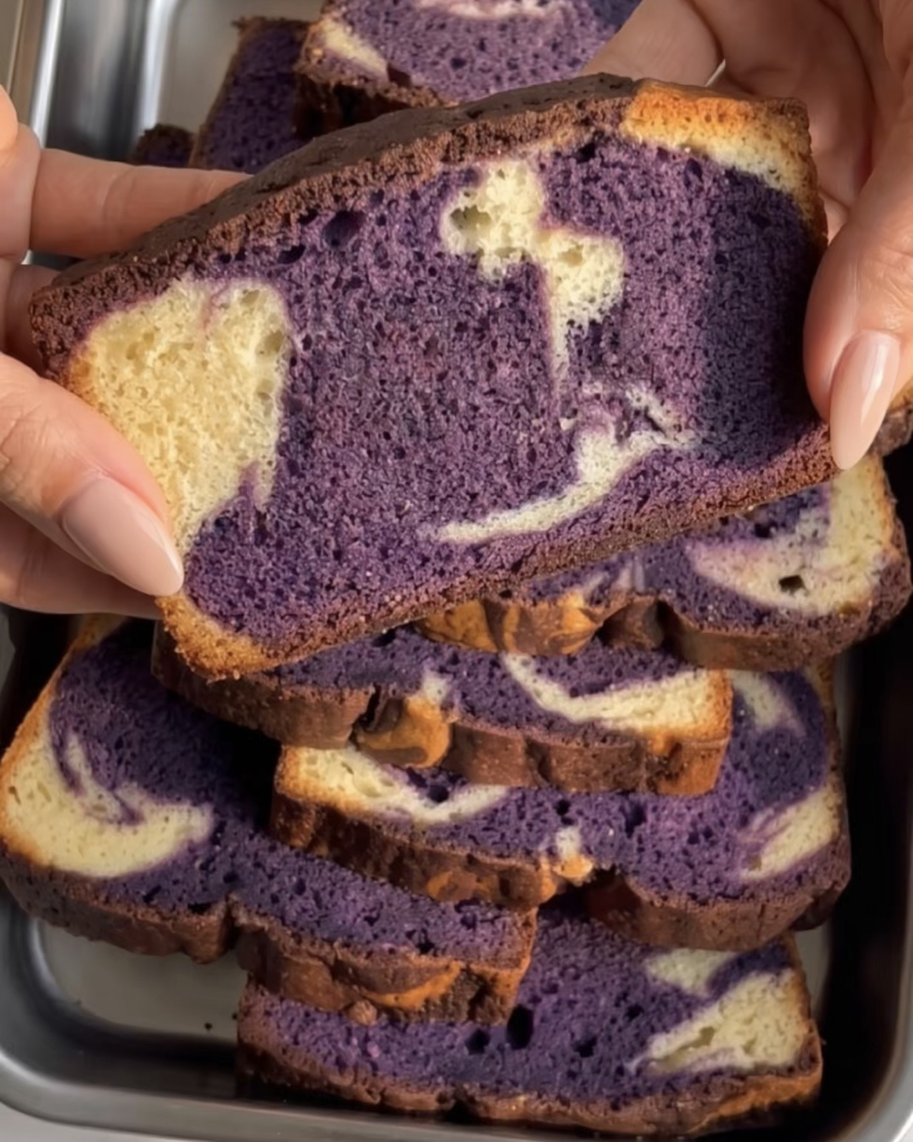 Mano sosteniendo una rebanada de bizcocho de ube marmoleado con un remolino morado y vainilla, horneado con polvo de ube UBE NOLIA®