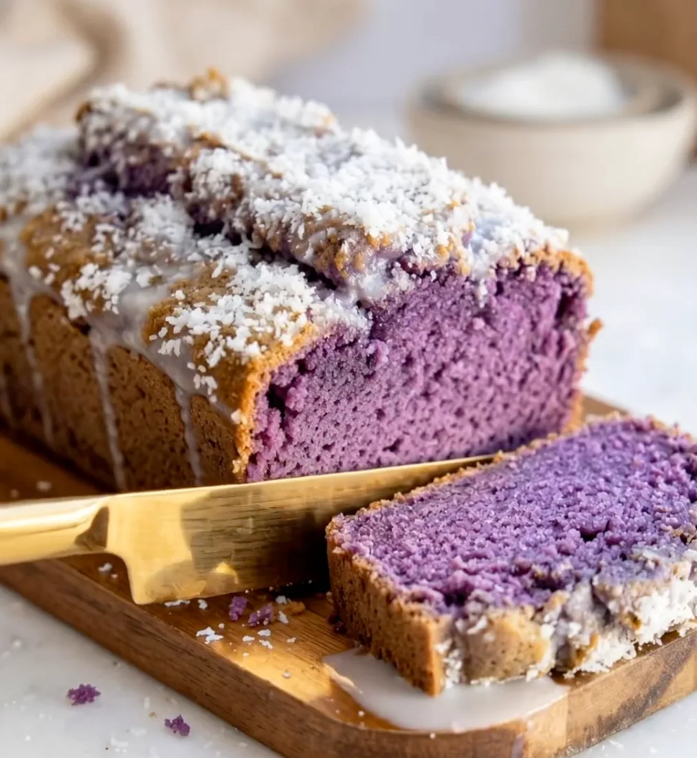 Bizcocho de ube y coco morado en rodajas sobre una tabla de madera con cobertura de coco blanco y un cuchillo dorado, receta Ube Nolia