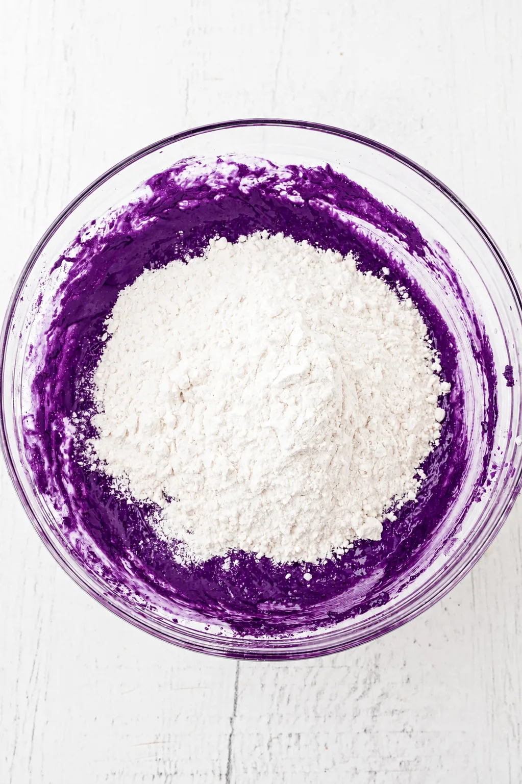 Harina añadida sobre la masa de brownie de ube y chocolate blanco morado intenso en un tazón de vidrio, lista para incorporar con polvo de ube Ube Nolia