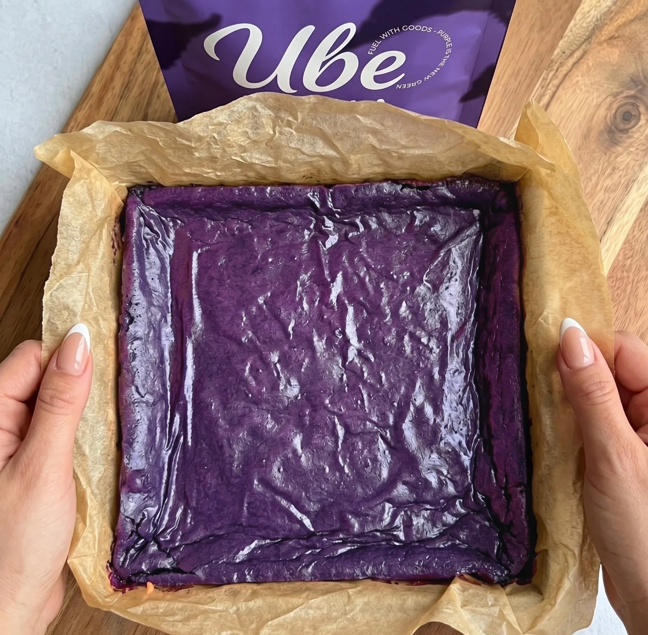 Masa de brownie de ube morado intenso en capas en un molde cuadrado forrado con papel de horno junto a un paquete de Ube Nolia powder